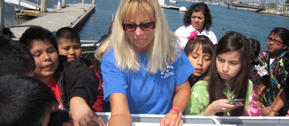 A woman in a blue shirt shows marine items to a group of children on a dock by the water.